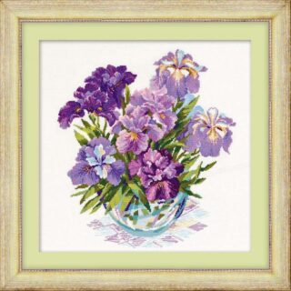 Cross-stitch embroidery of a vibrant bouquet of irises in shades of purple, lilac, and lavender, arranged in a glass vase with green leaves. The floral arrangement rests on a patterned cloth, framed in a light green mount with a wooden frame. Design from RIOLIS kit 1071.