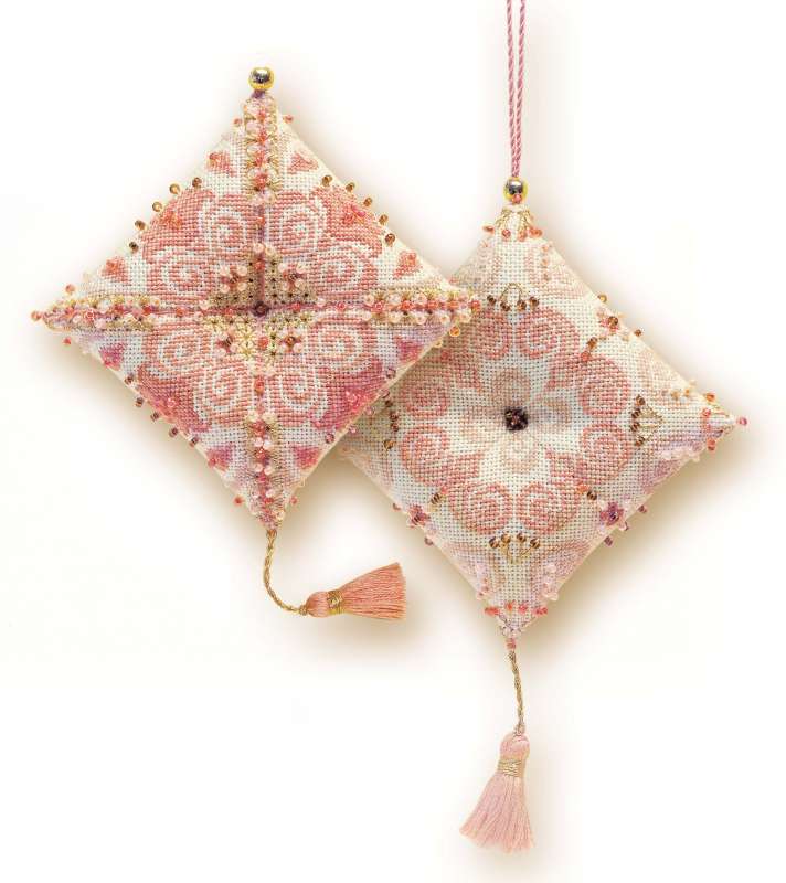 Cross-stitched diamond-shaped ornaments in pink and peach tones, featuring intricate patterns, gold beads, and pink tassels. Designed as hanging decorations or pincushions. Design from RIOLIS kit 1228 Zigouigoui Delight.