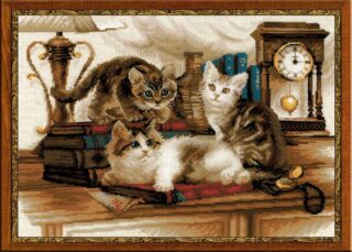 Cross-stitch embroidery of three playful kittens on a wooden desk, surrounded by books, a vintage lamp, and an antique clock. Two kittens sit upright while one lounges on its side. Design from RIOLIS kit 1247 Furry Friends.