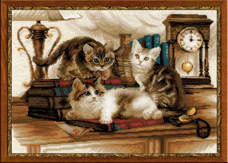 Cross-stitch embroidery of three playful kittens on a wooden desk, surrounded by books, a vintage lamp, and an antique clock. Two kittens sit upright while one lounges on its side. Design from RIOLIS kit 1247 Furry Friends.