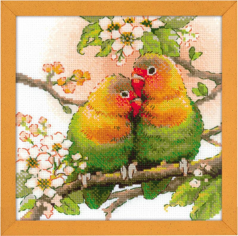 A cross-stitched pair of colorful lovebirds cuddling on a flowering tree branch with green leaves and white blossoms.