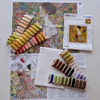 Klimt’s Lady with a Fan cross stitch shows a woman in a patterned robe holding a red fan surrounded by colorful birds and flowers.