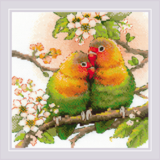 A cross-stitched pair of colorful lovebirds cuddling on a flowering tree branch with green leaves and white blossoms.