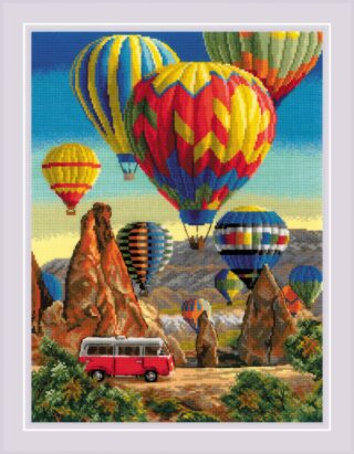 A cross-stitch scene of colorful hot air balloons floating above rocky cliffs in Cappadocia, with a red camper van driving below.
