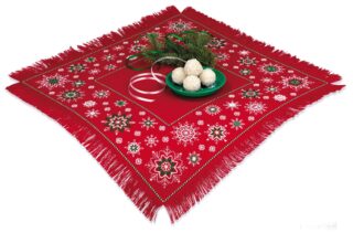 A red table topper with white and green embroidered snowflake patterns, fringed edges, and a festive holiday arrangement in the center.