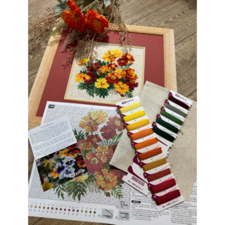 A cross-stitch artwork of bright marigolds in shades of yellow, orange, and red, arranged as a cheerful bouquet on cream fabric.