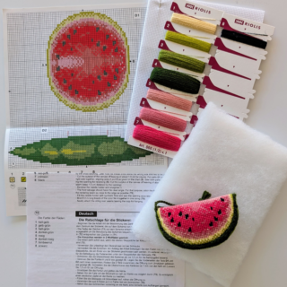 Cross-stitch design of a watermelon-shaped pincushion with pink flesh, green rind, and black seed details.