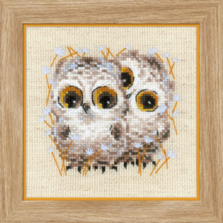 Cross-stitch of two baby owls with wide amber eyes, surrounded by delicate white flowers and orange stems on beige fabric.
