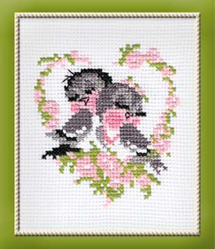 A sweet cross-stitch design of two young birds nestled together inside a heart-shaped frame of pink flowers and greenery.