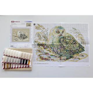 Cross-stitch embroidery of a white owl with golden eyes, resting among snow-covered grasses. The owl’s feathers are detailed in white, grey, and black tones. Design from RIOLIS kit 1241 White Owl.