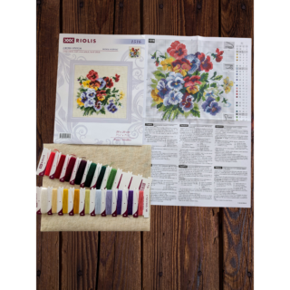 Cross-stitch artwork of a vibrant bouquet featuring red, yellow, purple, and white pansies arranged against a neutral fabric background, framed with an elegant beige and gold frame.