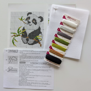 Cross-stitch of a cute panda holding bamboo leaves, stitched in black, white, and green, framed in red.