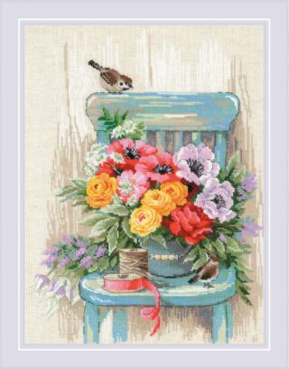 Cross-stitch of a bouquet of colorful garden flowers arranged on a teal chair with two small birds, a spool of thread, and ribbon beside it.