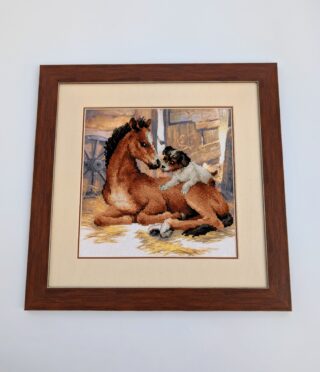 Framed cross stitch artwork of a foal lying on hay in a stable with a playful puppy climbing onto its back.