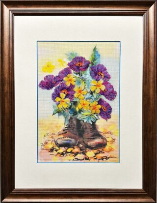 Framed cross stitch artwork featuring worn leather boots repurposed as planters, filled with blooming purple and yellow flowers.