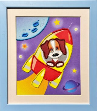 Framed embroidery of a cartoon puppy riding in a red and yellow rocket flying through space with stars, the moon, and a planet.