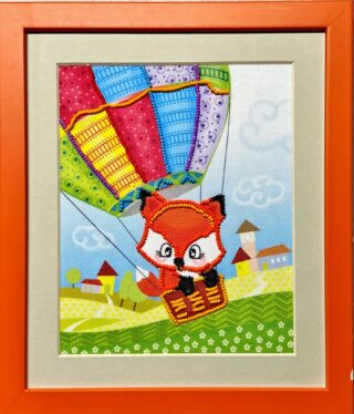 Framed embroidery of a cartoon fox sitting in a hot air balloon basket, floating above a colorful countryside village.