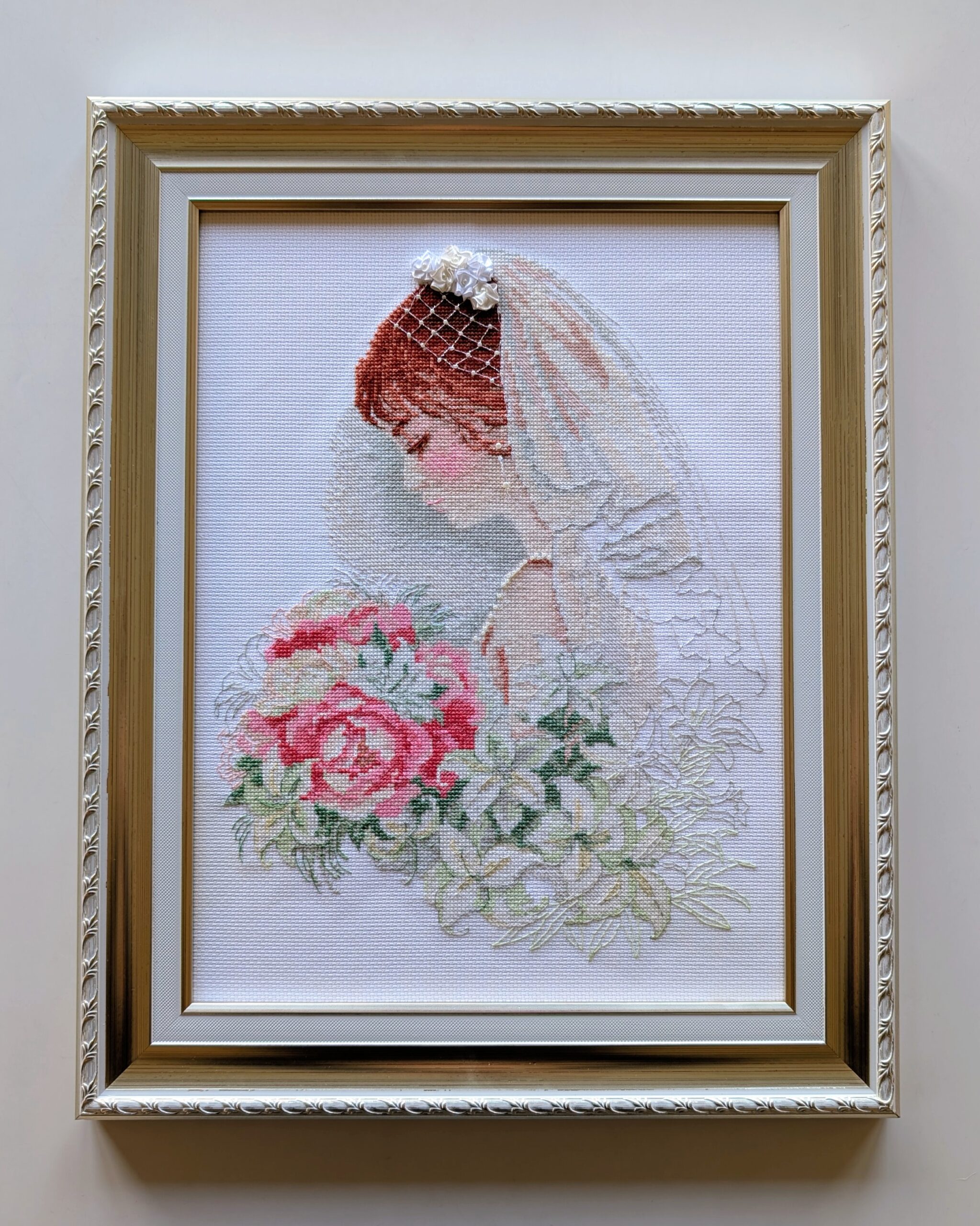 Framed embroidery of a bride in a white veil with flowers in her hair, holding a bouquet of pink and white blossoms.
