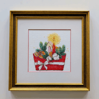 Framed cross stitch artwork of a festive red envelope with greenery, dried oranges, cotton, and a glowing candle, part of exhibition models.