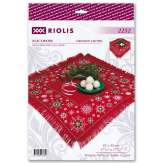 A red table topper with white and green embroidered snowflake patterns, fringed edges, and a festive holiday arrangement in the center.