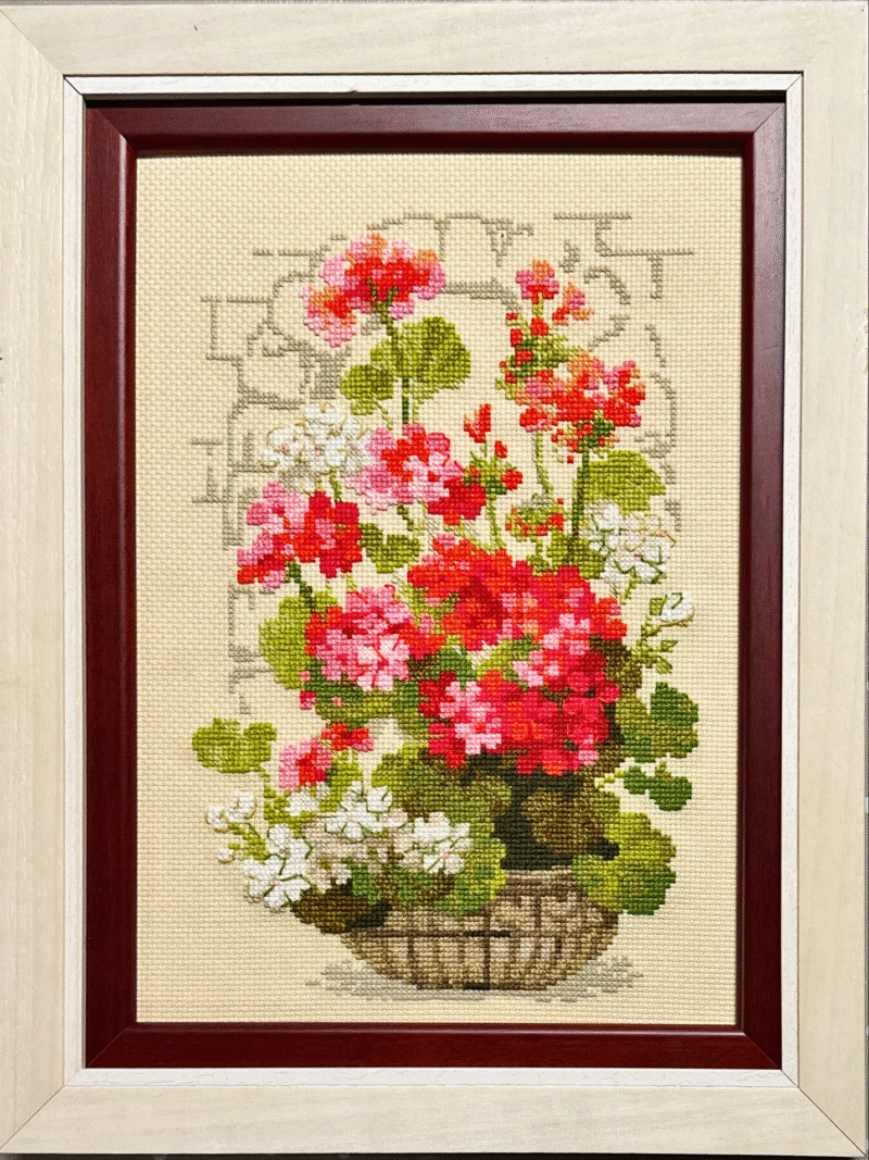 Framed embroidery of a wicker basket filled with red, pink, and white geraniums stitched on beige fabric.