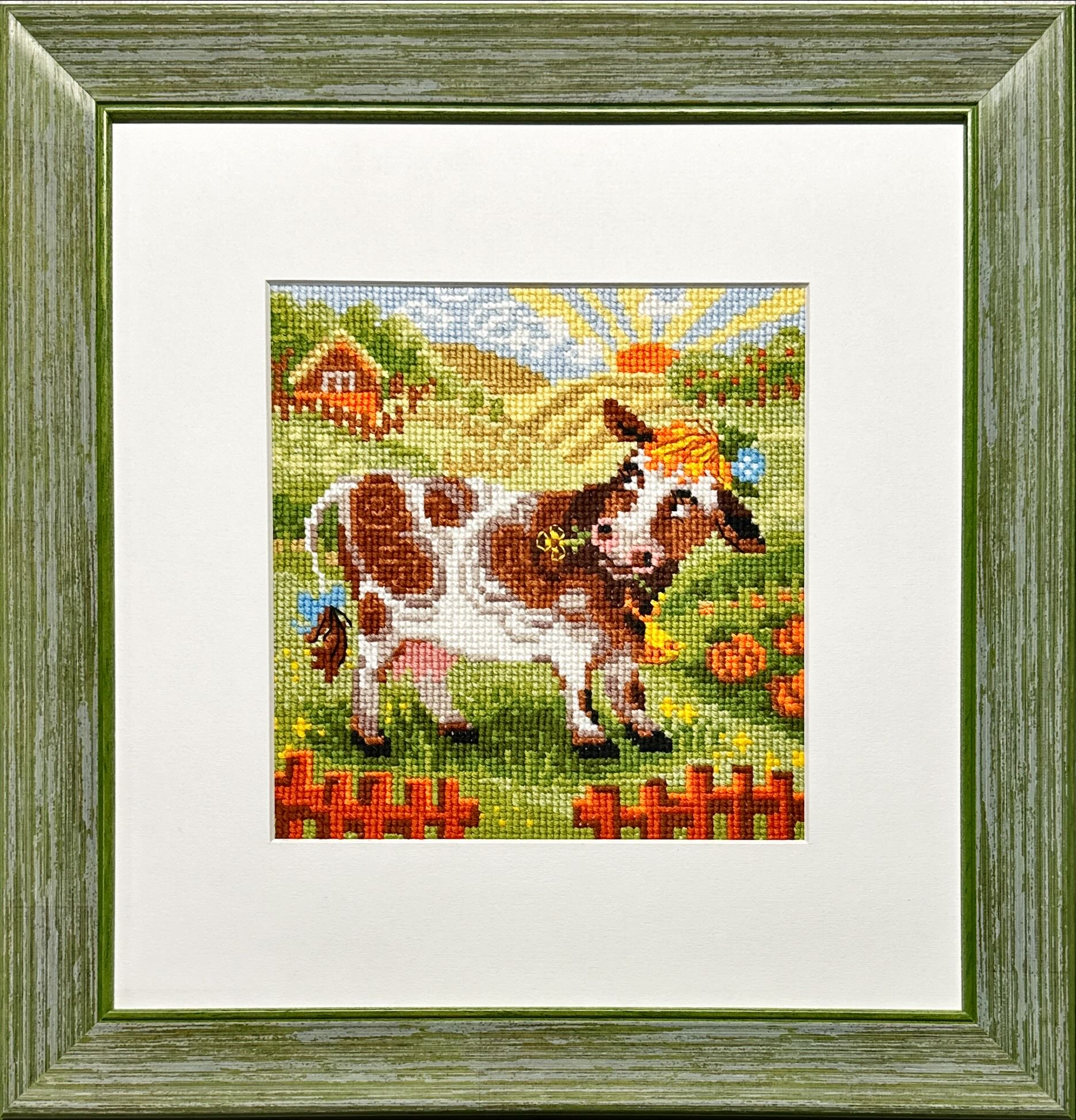 Framed embroidery of a brown-and-white cow standing in a sunny meadow with flowers, pumpkins, and a farmhouse in the background.