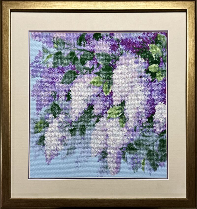 Framed embroidery of lilac flowers in shades of purple and white with green leaves on a blue background.