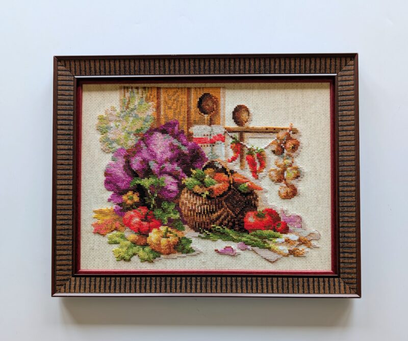 Framed embroidery of a rustic still life with a basket of vegetables, garlic, and peppers in a warm kitchen setting.