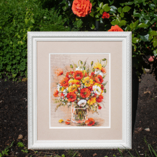 A vibrant cross-stitched bouquet of red, yellow, and white field poppies arranged in a glass vase, with scattered petals around the base.