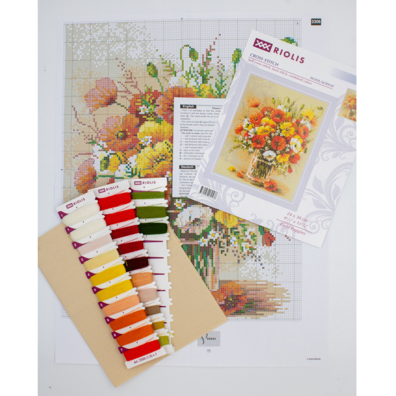 A vibrant cross-stitched bouquet of red, yellow, and white field poppies arranged in a glass vase, with scattered petals around the base.