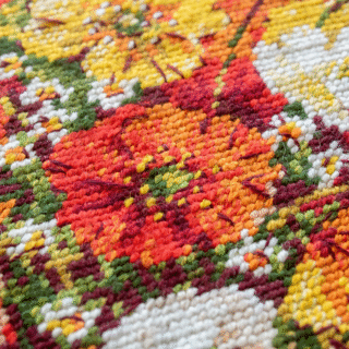 A vibrant cross-stitched bouquet of red, yellow, and white field poppies arranged in a glass vase, with scattered petals around the base.