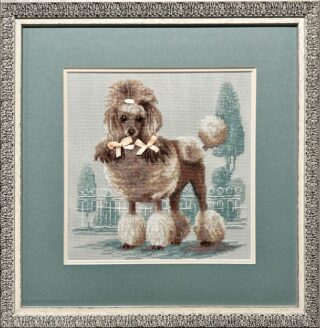 Framed cross-stitch artwork featuring a well-groomed poodle with bows, standing in front of a soft, pastel garden background.