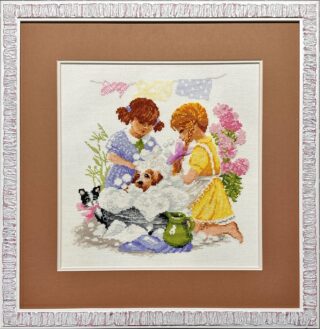 Framed cross-stitch artwork showing two girls washing a small puppy in a tub filled with bubbles, surrounded by flowers and playful details.