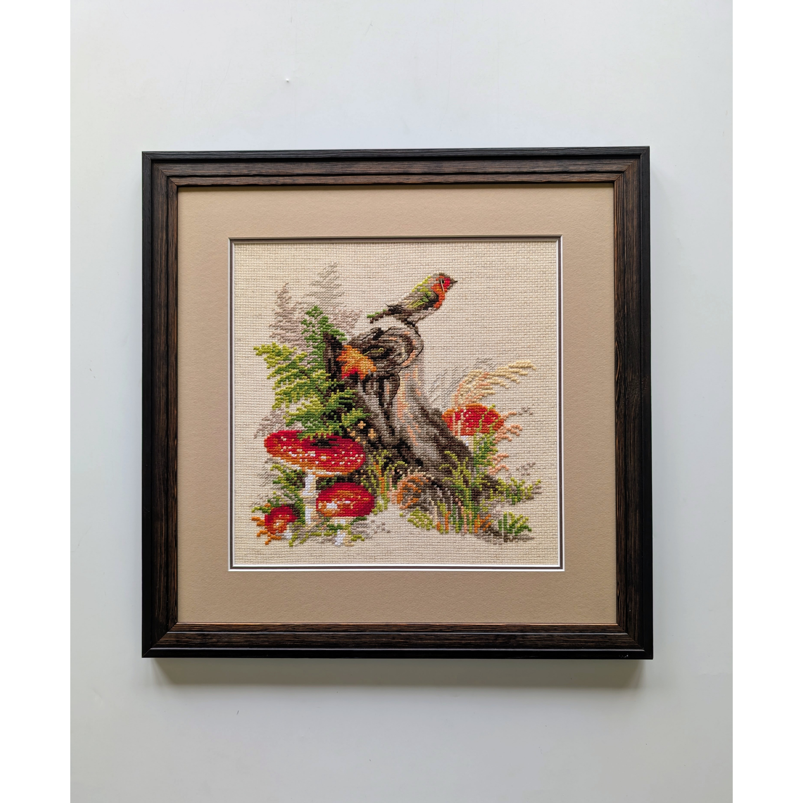 Framed cross-stitch artwork featuring a colorful bird perched on a tree stump surrounded by bright red mushrooms, foliage, and forest grasses.