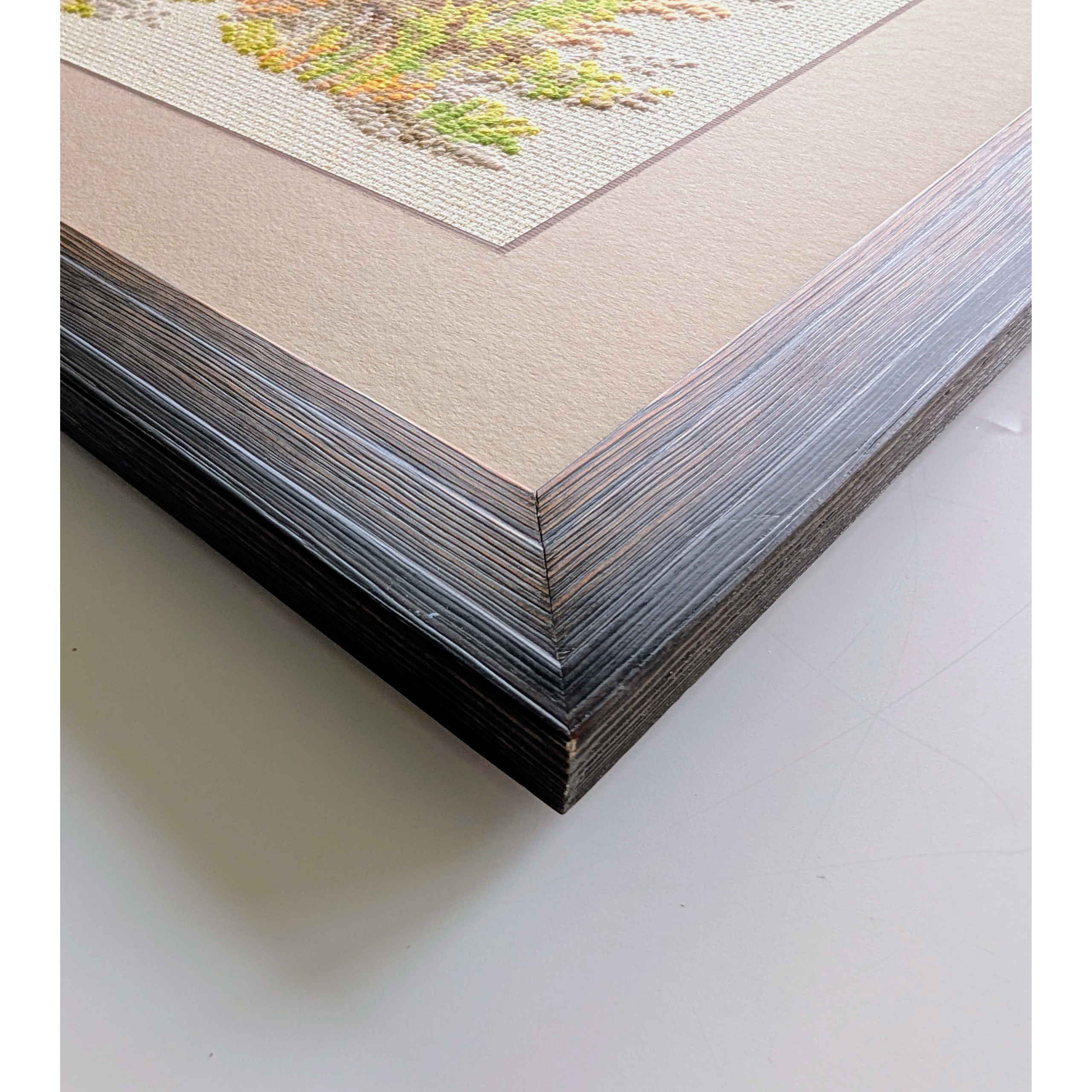 Framed cross-stitch artwork featuring a colorful bird perched on a tree stump surrounded by bright red mushrooms, foliage, and forest grasses.