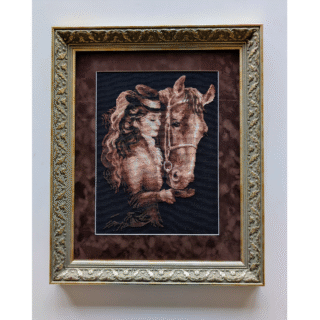 Framed cross-stitch artwork depicting a woman in vintage attire standing closely beside a horse on a dark background.
