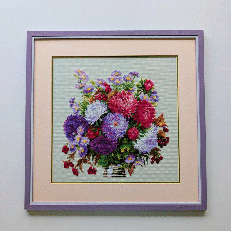 Framed cross-stitch artwork featuring a large colorful bouquet of asters in shades of purple, pink, red, and white.