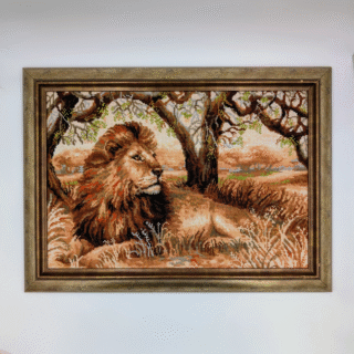 Framed cross-stitch artwork showing a majestic lion resting under trees in a warm savanna landscape.