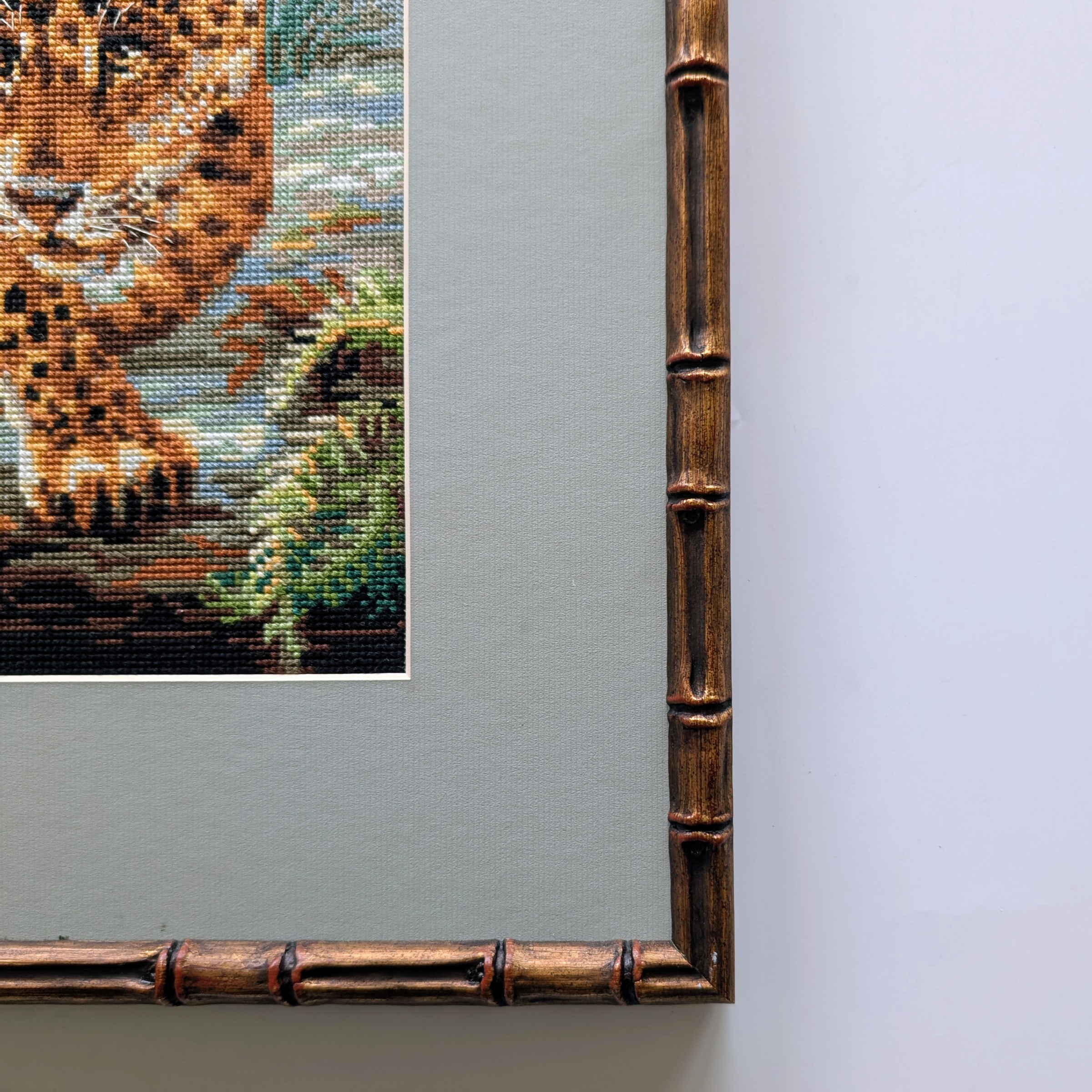 Framed cross-stitch artwork depicting a leopard stepping forward on jungle rocks, surrounded by lush green foliage.