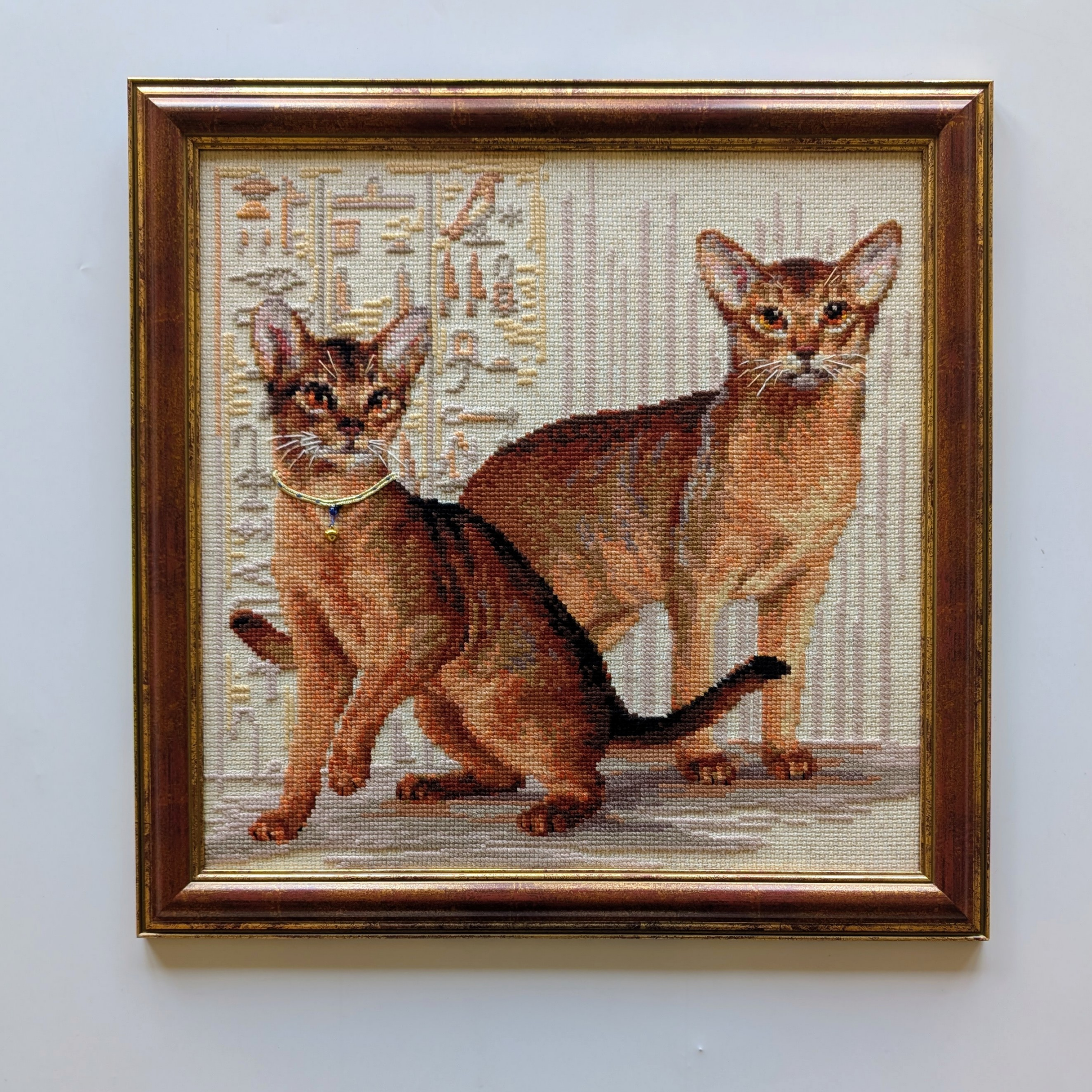 Framed cross-stitch artwork showing two Abyssinian cats posed against a background with Egyptian-style patterns.