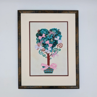 Framed cross-stitch artwork of a heart-shaped tree with decorative leaves, swirling branches, and pastel bows in a patterned pot.