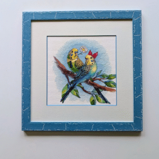 Framed cross-stitch artwork of two colorful budgerigars perched on a branch, one holding a daisy and the other wearing a red bow.