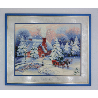 Framed cross-stitch artwork showing a snowy winter scene with a cozy house, pine trees, a small bridge, and a horse-drawn sleigh at Christmas time.