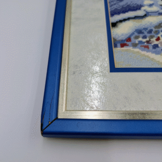 Framed cross-stitch artwork showing a snowy winter scene with a cozy house, pine trees, a small bridge, and a horse-drawn sleigh at Christmas time.