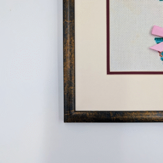 Framed cross-stitch artwork of a heart-shaped tree with decorative leaves, swirling branches, and pastel bows in a patterned pot.