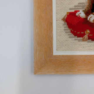 Framed cross-stitch artwork of a fluffy brown and cream cat sitting on a red cushion against a neutral background.