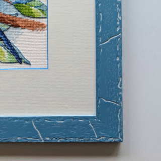 Framed cross-stitch artwork of two colorful budgerigars perched on a branch, one holding a daisy and the other wearing a red bow.