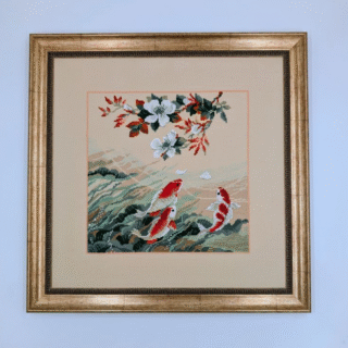Framed cross-stitch artwork showing red and white koi fish swimming among flowing waves beneath flowering branches.