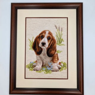 Framed cross-stitch artwork depicting a basset hound puppy sitting on grass with small blue flowers and tall meadow plants behind it.