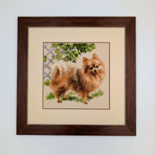 Framed cross-stitch artwork showing a fluffy Pomeranian dog standing in a garden with green foliage and a trellis in the background.
